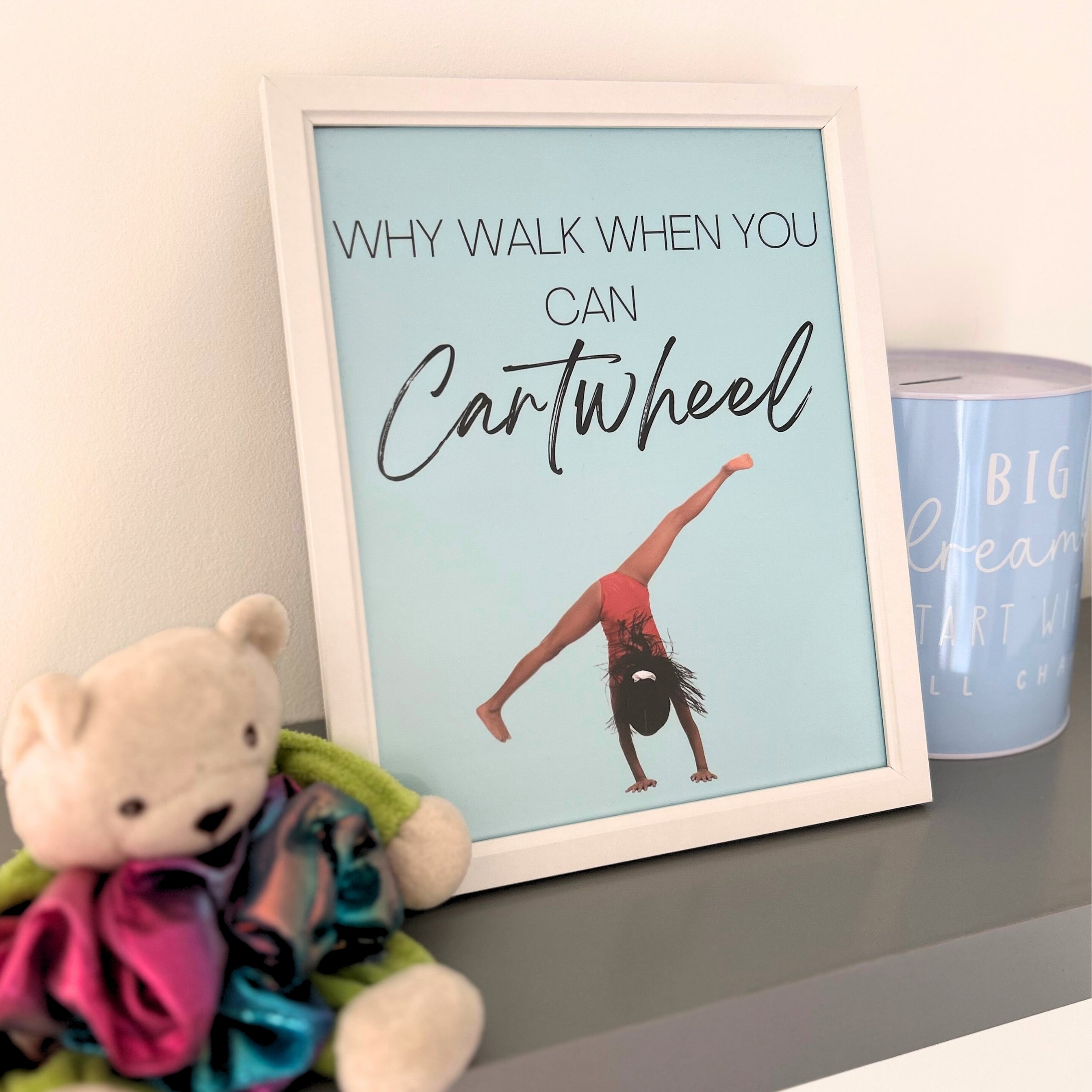 Framed gymnastics print with a girl doing a cartwheel and text 'Why walk when you can cartwheel' on a shelf with a teddy bear and mug.