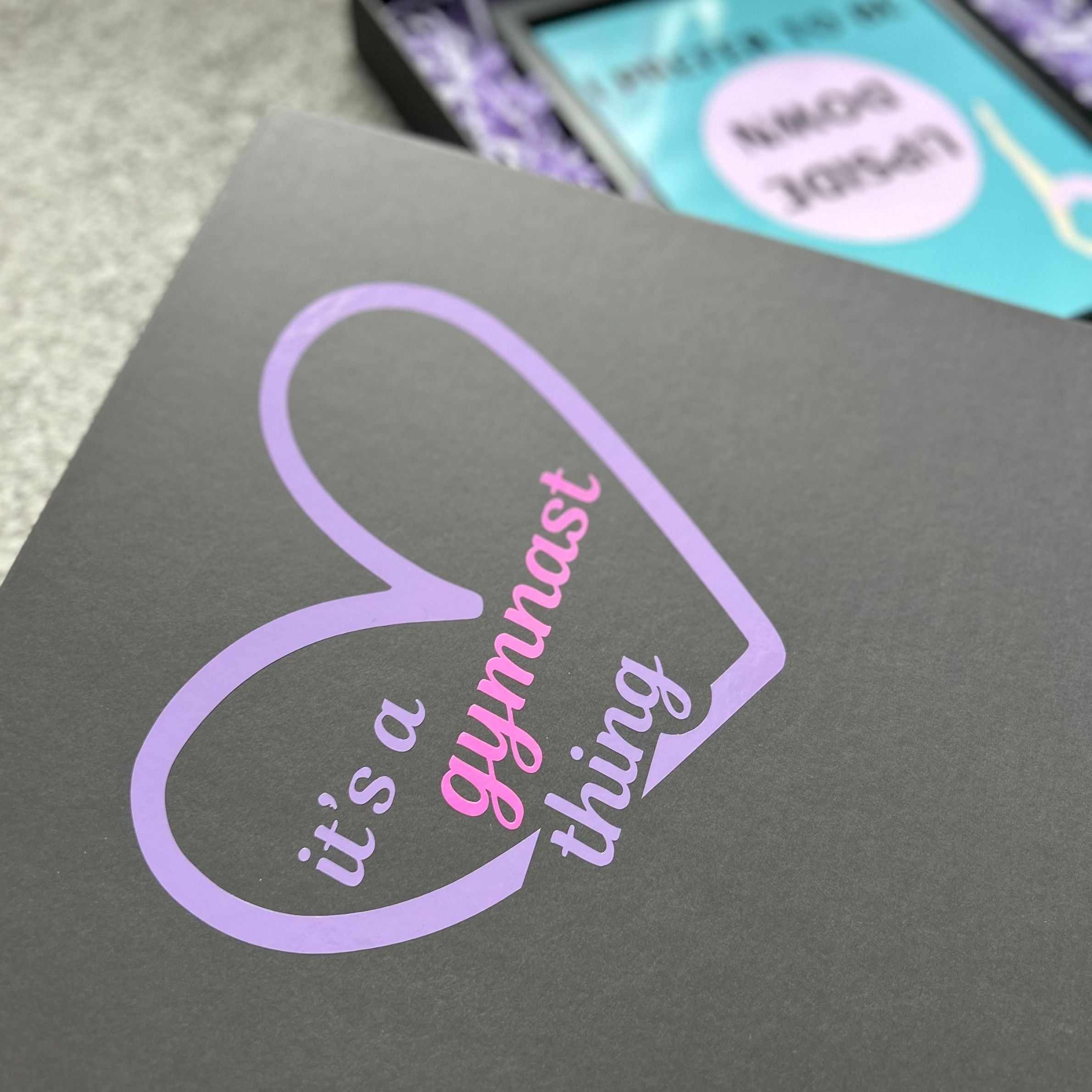 Grey gymnastics gift box with a purple heart and pink text 'it's a gymnast thing' on a textured surface.