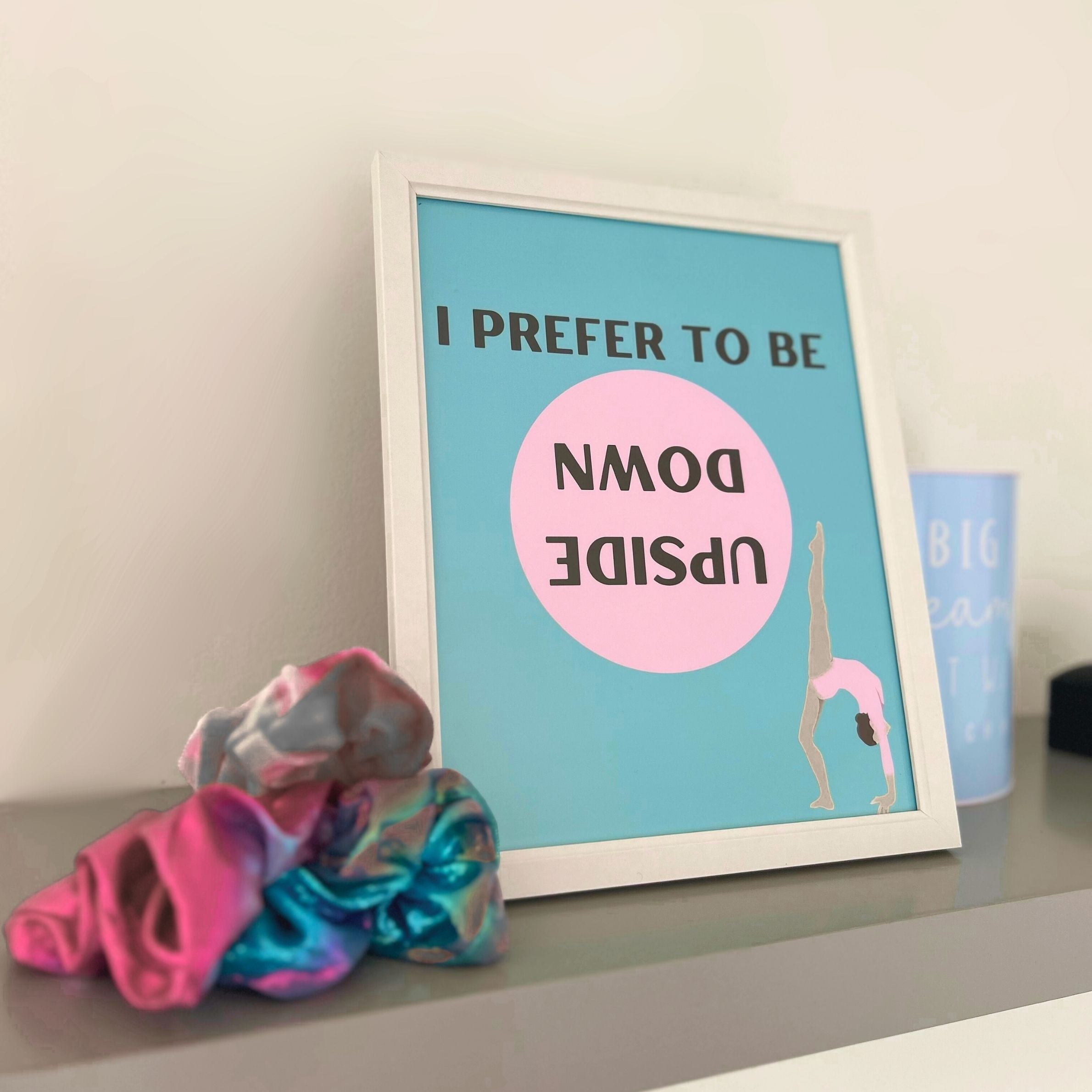 Framed poster for gymnast with text and illustration of a gymnast upside down, next to colourful scrunchies on a surface.