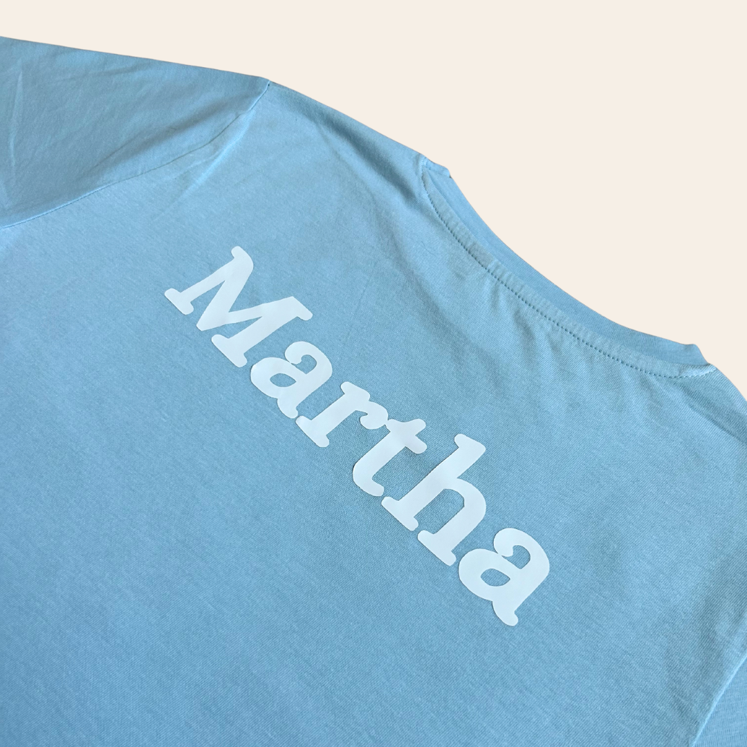 Light blue personalised gymnastics top with 'Martha' printed on it against a beige background