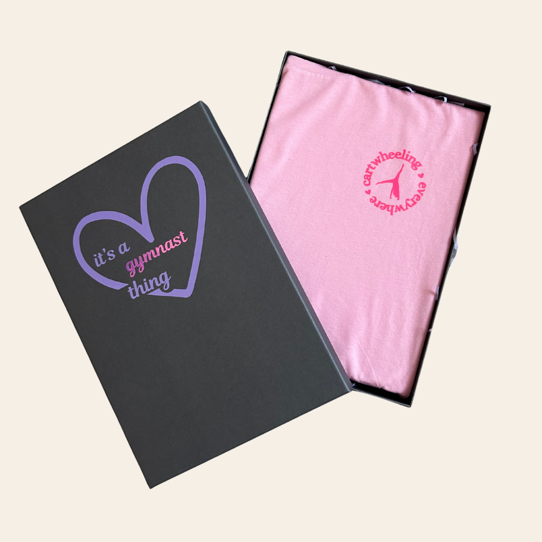 Grey gymnastics gift box with purple text reading 'its a gymnast thing' and pink gymnastics t-shirt with cartwheel logo on a beige background