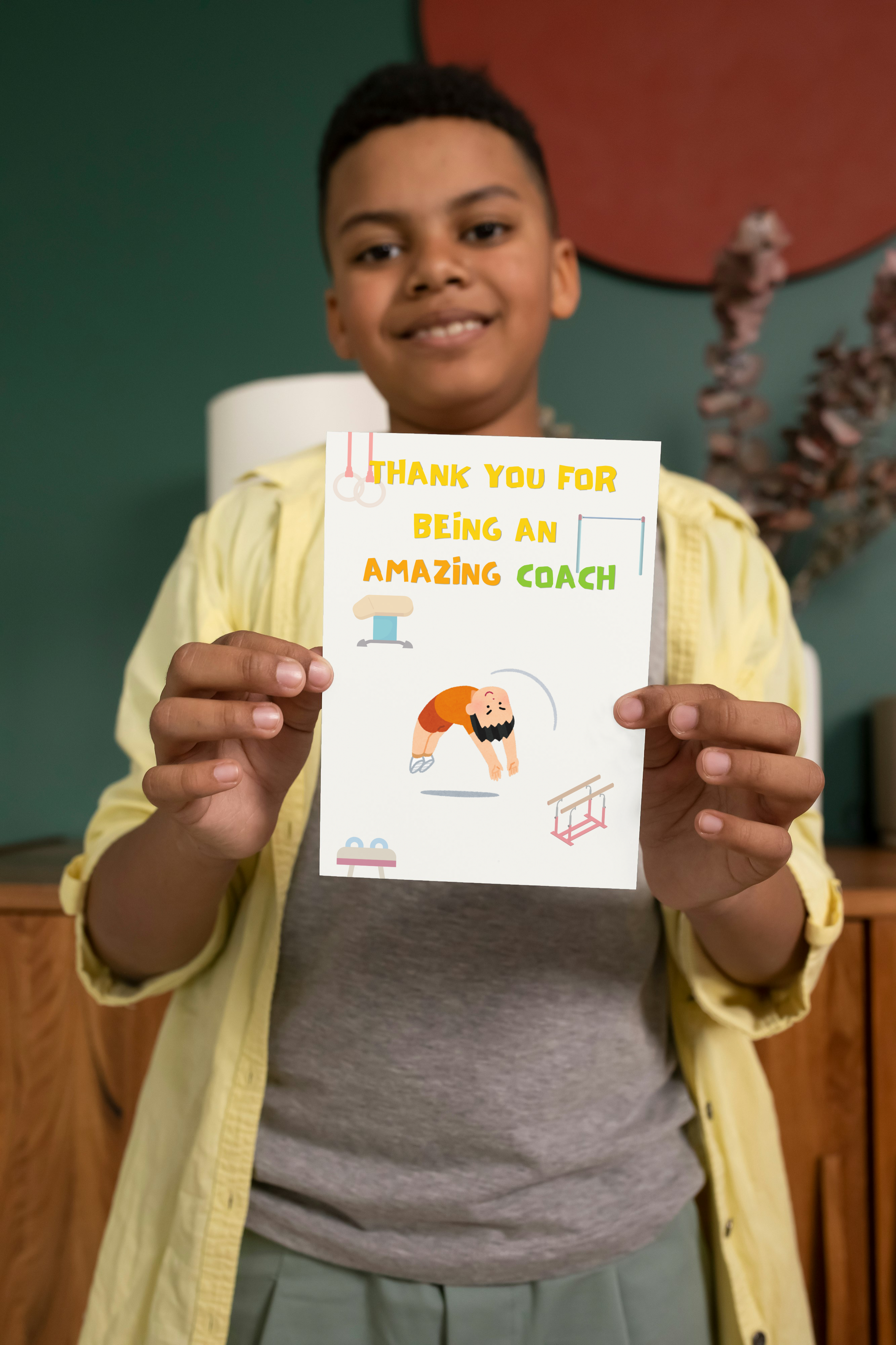 Boy holding a thank you card for gymnastics coach that says 'Thank you for being an amazing coach' with a cartoon illustration.
