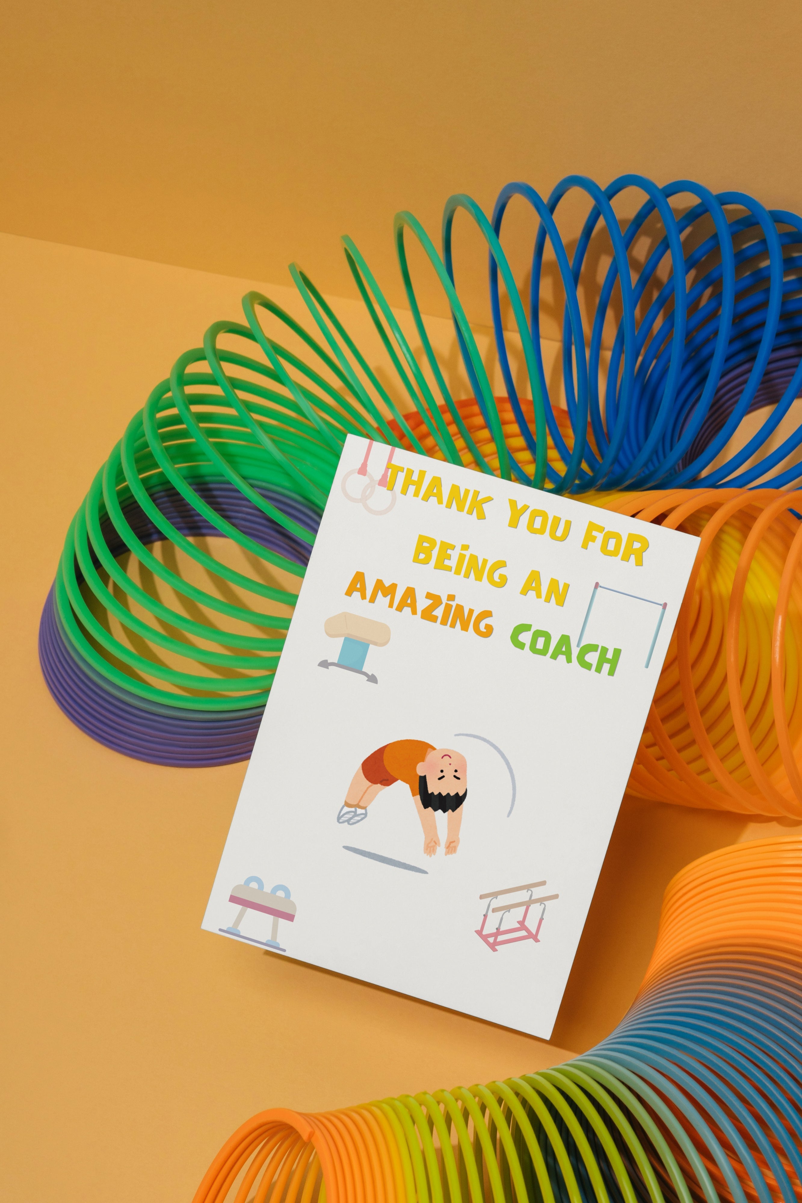Colourful slinky toy with a 'Thank you for being an amazing coach' gymnastics coach thank you card on a yellow background