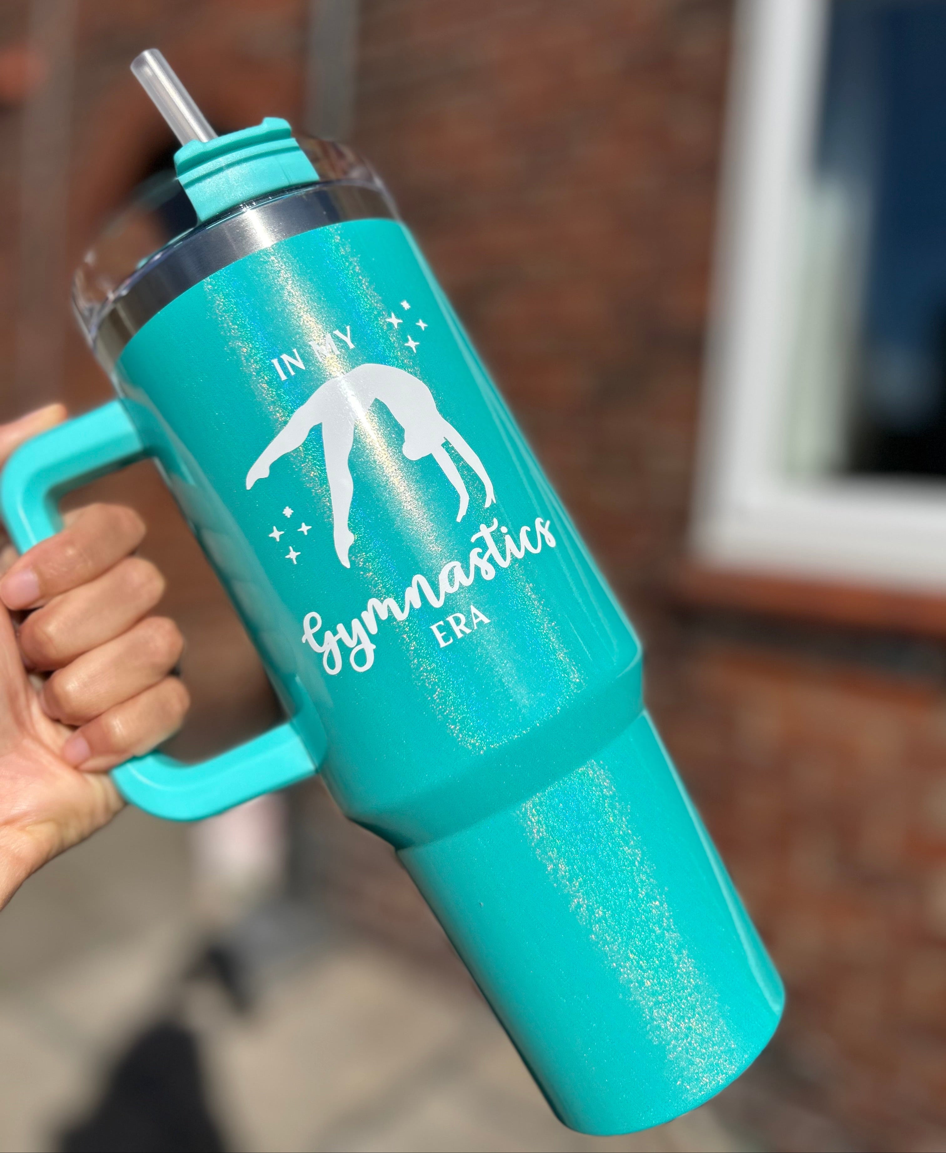 Turquoise gymnastics stanley dupe tumbler with handle held by a hand, featuring a gymnast design and text.